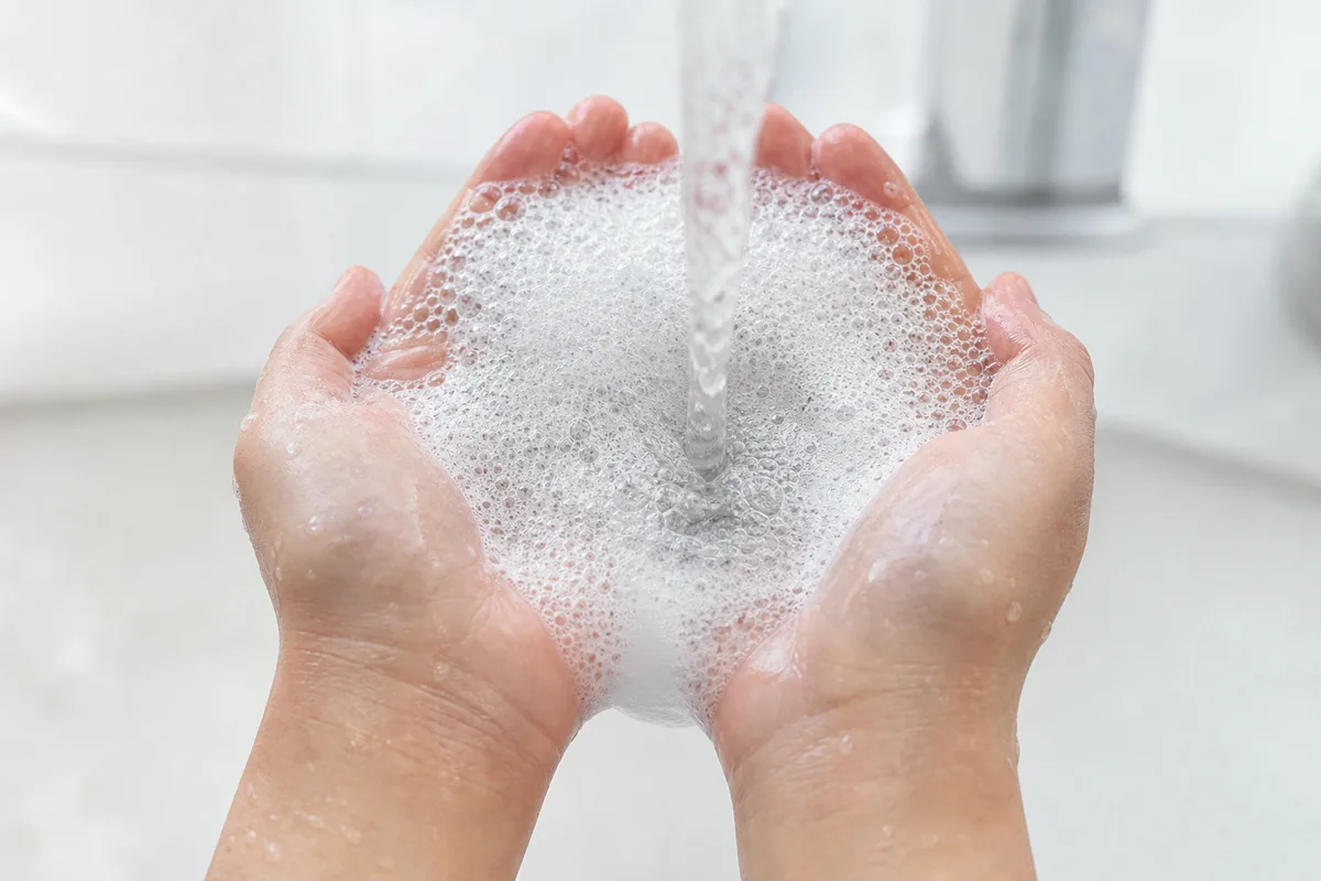 Mousse de shampooing blanche dans deux mains en coupe sous un filet d’eau